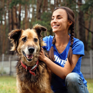 young brunette woman wearing a volunteer t-shirt petting a mixed breed, fuzzy, beige and black, 40lb dog with a red collar